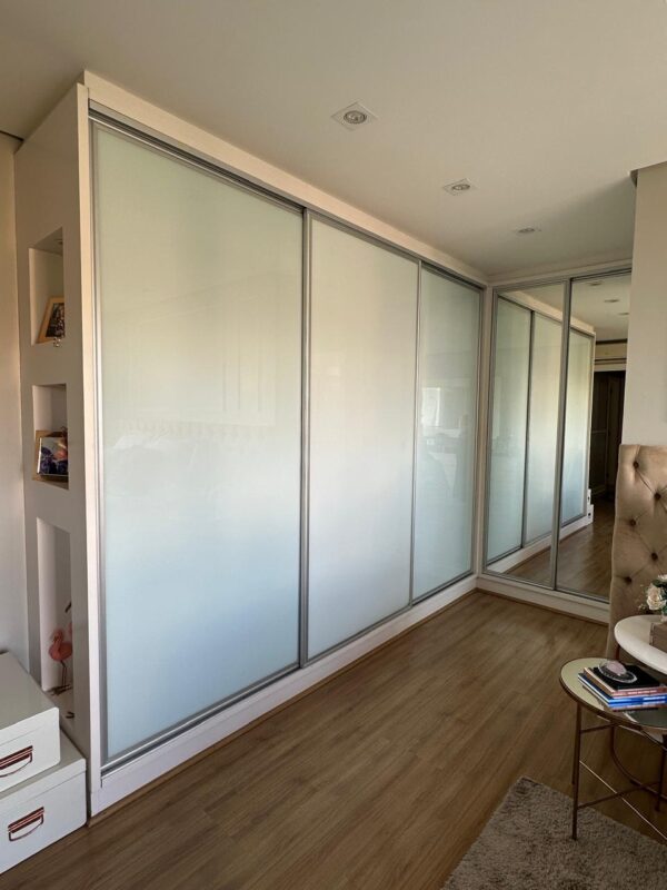 ARMÁRIO EM “L” — PORTAS DE CORRER — 3 DELAS DE VIDRO SERIGRAFADO + 2 DELAS EM ESPELHO — PARTE INTERNA COMPOSTA POR GAVETAS, PRATELEIRAS, ARARAS, GAVETAS COM DIVISÓRIAS PARA BIJUS, CALCEIROS, ÁREA PARA ROUPAS LONGAS — MDF COM LACA BRANCA — NICHOS LATERAIS COM ILUMINAÇÃO